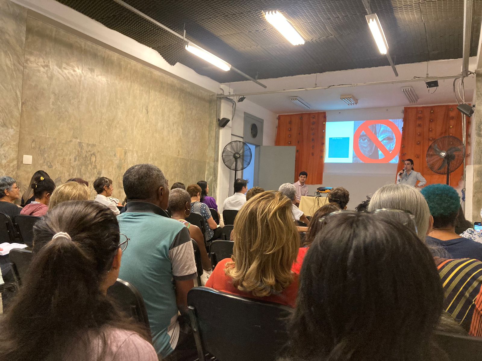 Palestrante Eduarda durante resumo do que é etarismo. No espaço estão atendidos e colaboradores do serviço. As paredes do espaço sao marmorizadas, e no palco há duas cortinas laranja.